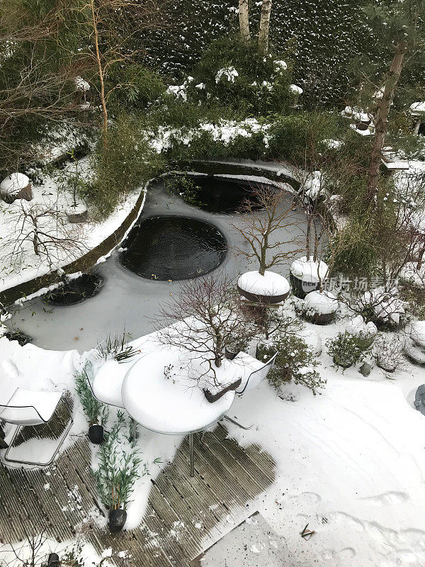 大冰封的锦鲤池在冬天的花园被冰雪覆盖后，大雪/暴风雪，金鱼池泵与曝气气泡阻止水冻结的东方日本园林盆景树和禅宗灯笼